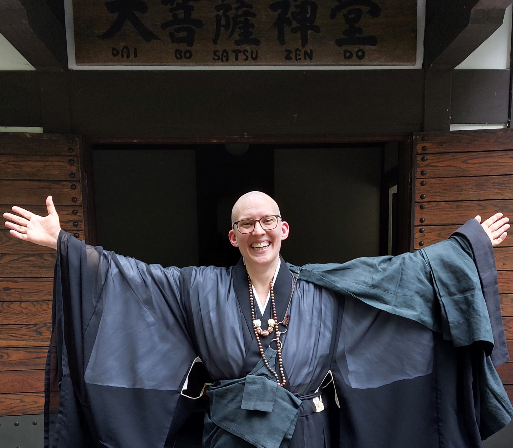 Ich stehe mit ausgebreiteten Armen und in einer Zen Priesterrobe vor dem Eingang des Dai Bosatsu Zendo