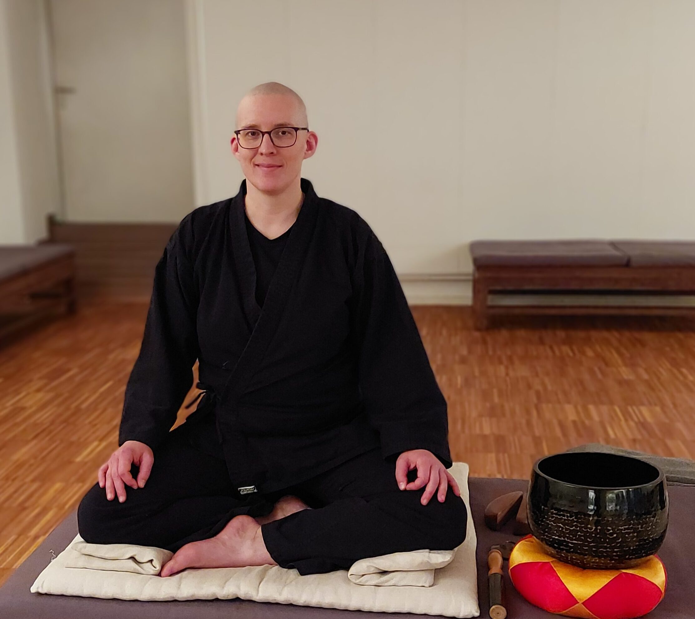 Ich sitze in Meditationskleidung in einem Zendo auf einem Podest mit einer Klangschale neben mir und lächle in die Kamera.