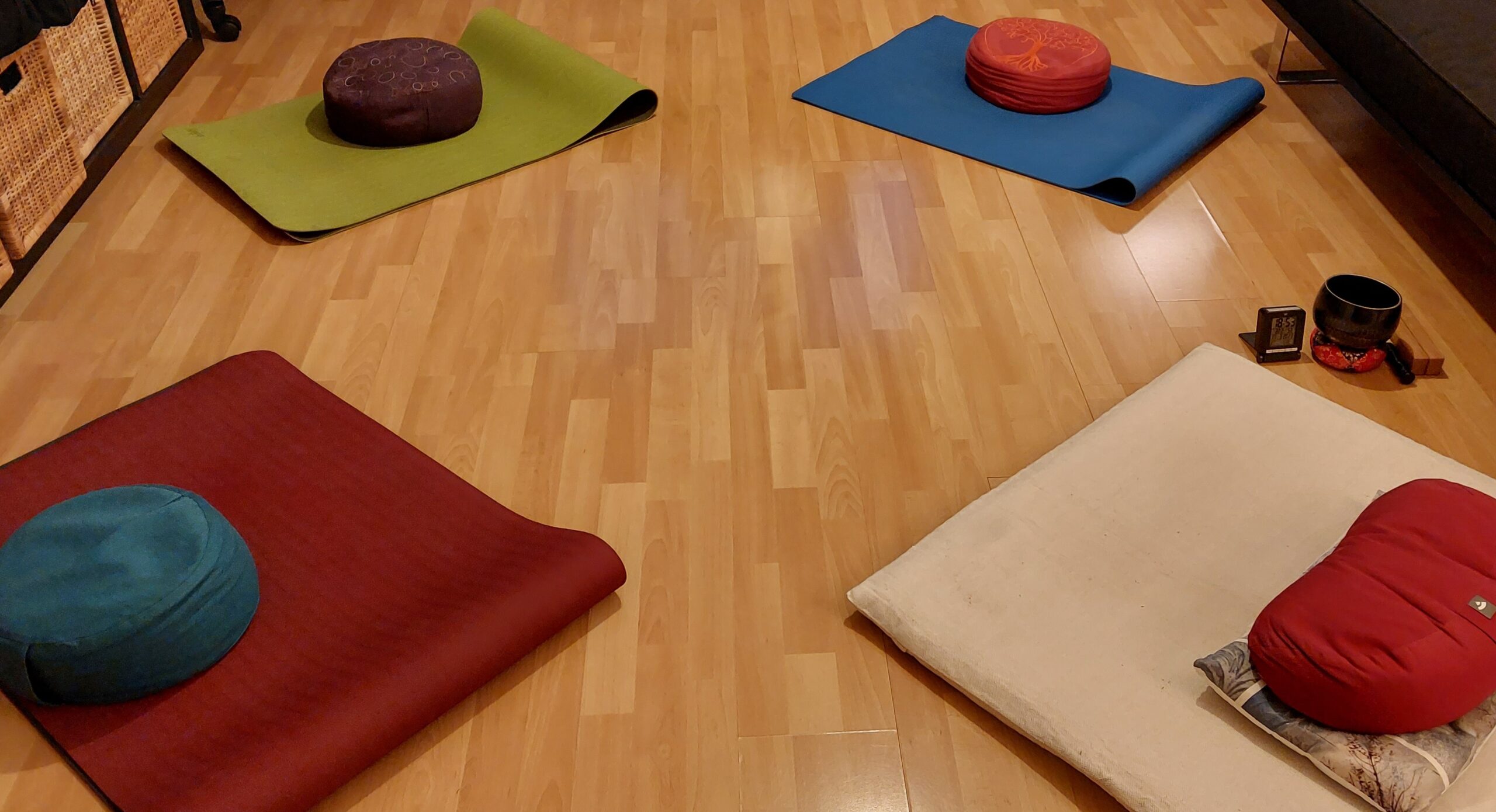 Vier Meditationskissen und vier Unterlagen, alles in verschiedenen Farben liegen im Quadrat zueinander auf einem Parketboden verteilt.