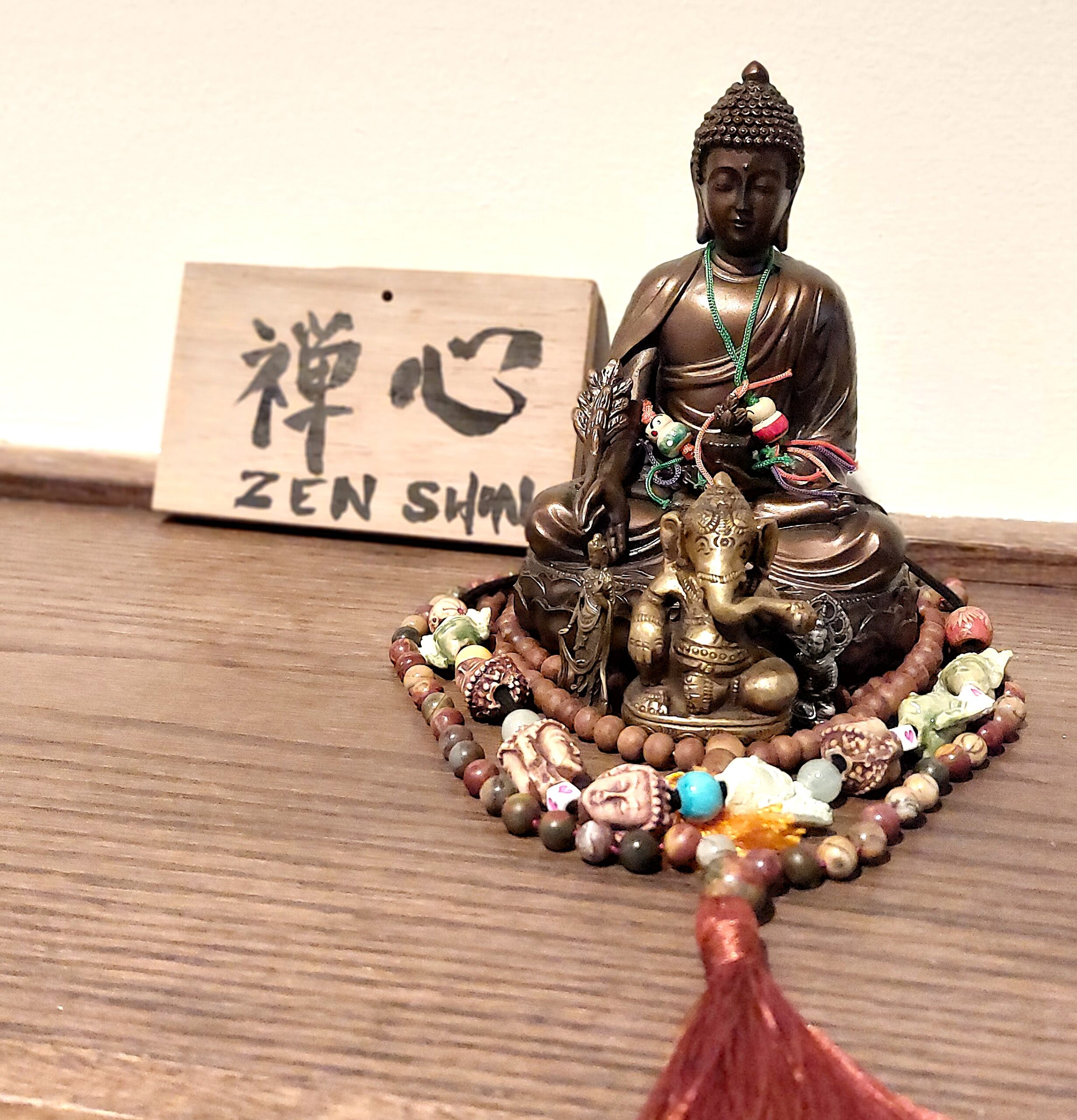 Ein Schild mit dem Namen Zenshin in Kanji und Romanji steht schräg hinter einer zwei Hände großen sitzenden Buddhastatue, die noch mit diversen Mala und einer weiteren Figur "geschmückt" ist